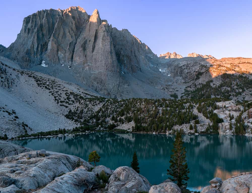 Mountain cliff overlooking a shaded lake. Sunrise is lighting up the top of the mountain.