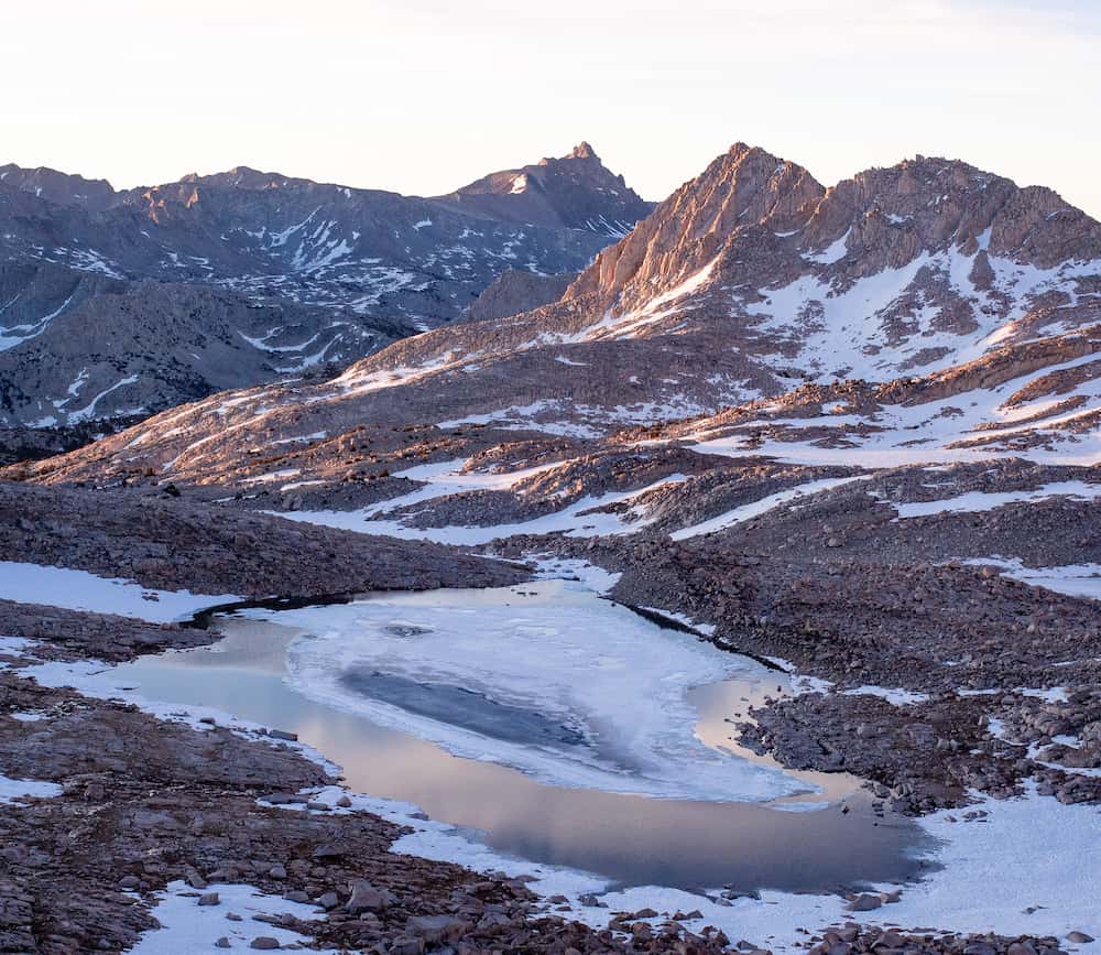 Semi-frozen lake surouned by rocks. Peaks visible in the distance, lit by the sunrise.