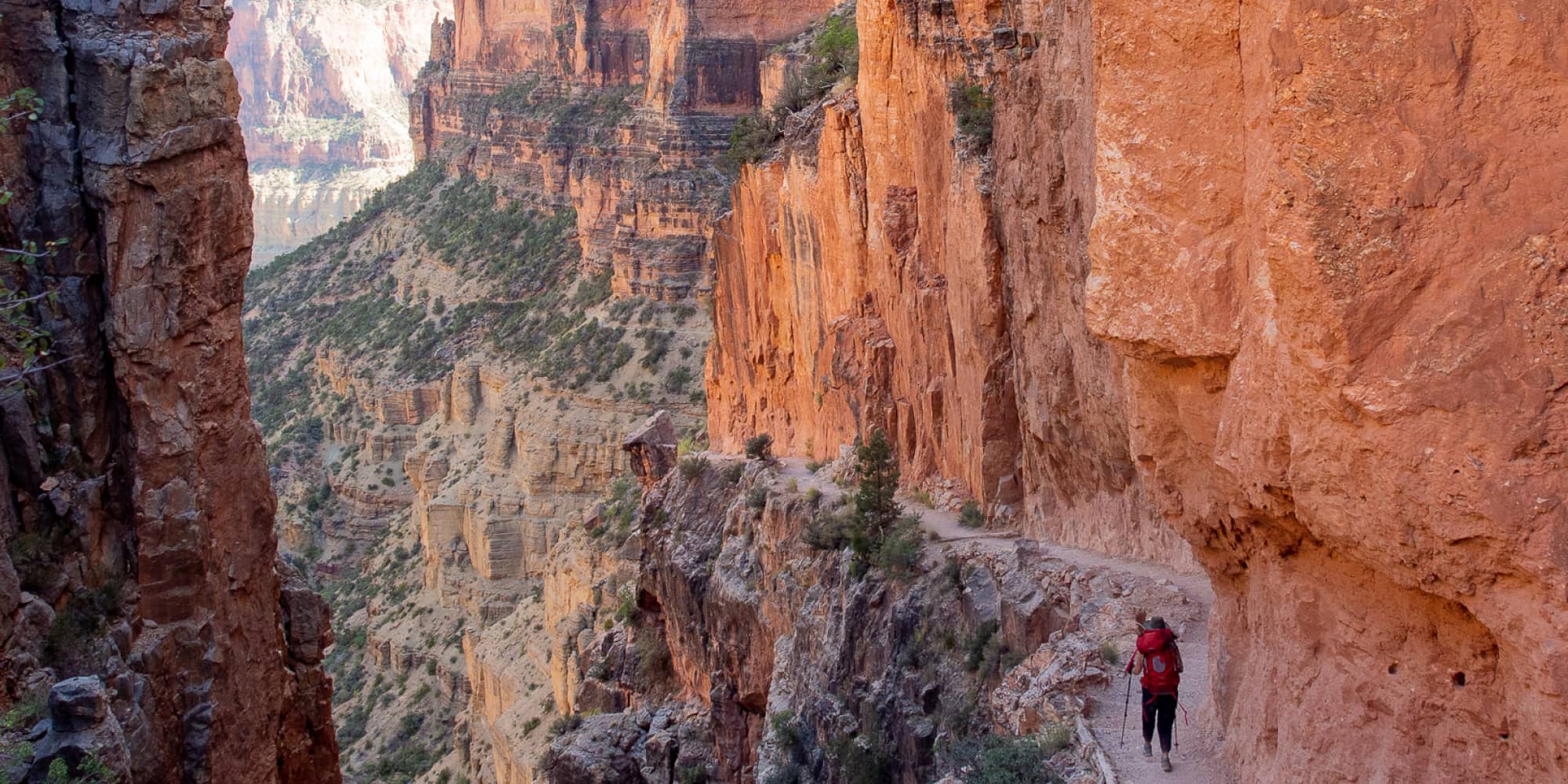 A backpacker hiking in the Grand Canyon. They are surounded by orange cliffs. A path is carved in the side of the cliff on which the backpacker is traveling.